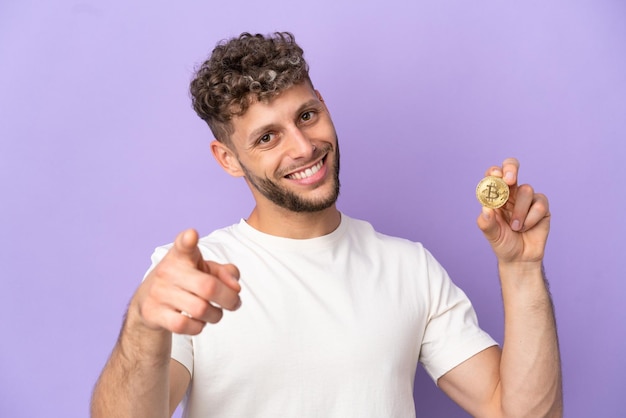 Jonge blanke man met een Bitcoin geïsoleerd op een paarse achtergrond die naar voren wijst met een gelukkige uitdrukking