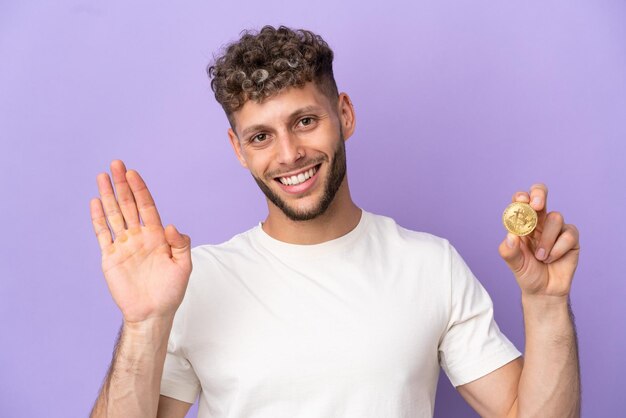 Jonge blanke man met een Bitcoin geïsoleerd op een paarse achtergrond saluerend met de hand met een gelukkige uitdrukking