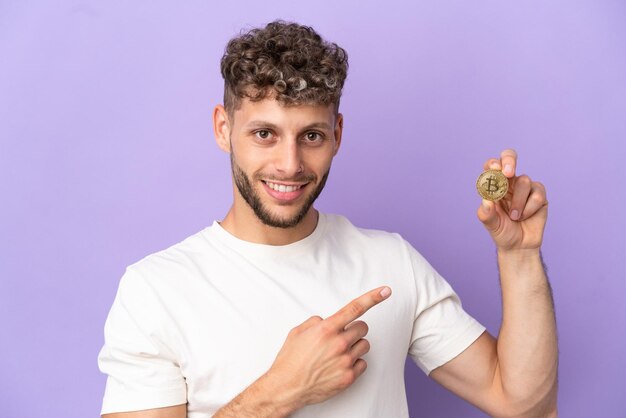 Jonge blanke man met een Bitcoin geïsoleerd op een paarse achtergrond wijzende vinger naar de zijkant