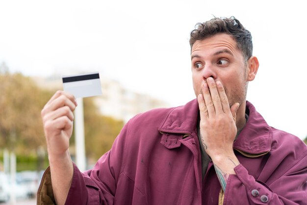 Jonge blanke man met een creditcard buitenshuis met verbazing en geschokte gezichtsuitdrukking