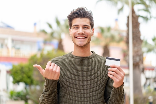 Jonge blanke man met een creditcard buitenshuis wijzend naar de zijkant om een product te presenteren