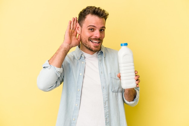 Jonge blanke man met een fles melk geïsoleerd op een gele achtergrond die probeert te luisteren naar een roddel.