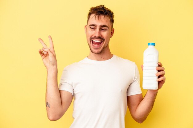 Jonge blanke man met een fles mil geïsoleerd op gele achtergrond vrolijk en zorgeloos met een vredessymbool met vingers.
