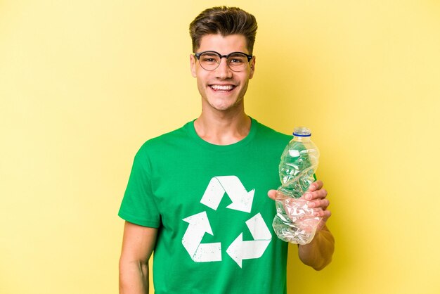 Jonge blanke man met een fles plastic om te recyclen geïsoleerd op gele achtergrond gelukkig, glimlachend en vrolijk.