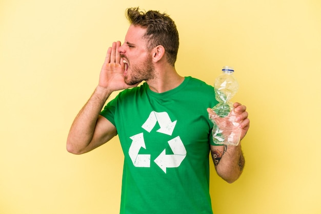 Jonge blanke man met een fles plastic om te recyclen geïsoleerd op gele achtergrond schreeuwen en palm in de buurt van geopende mond te houden.