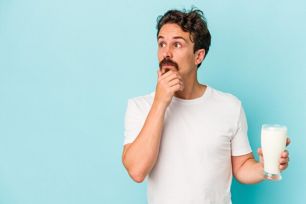 Jonge blanke man met een glas melk geïsoleerd op blauwe achtergrond zijwaarts kijkend met twijfelachtige en sceptische uitdrukking.