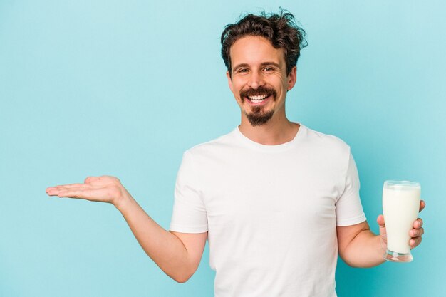 Jonge blanke man met een glas melk geïsoleerd op een blauwe achtergrond met een kopie ruimte op een palm en met een andere hand op de taille.