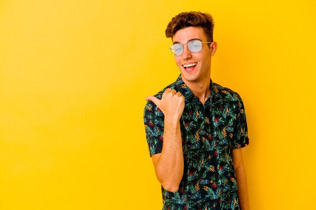 Jonge blanke man met een Hawaiiaans shirt geïsoleerd op gele achtergrond wijst met duimvinger weg, lachend en zorgeloos.