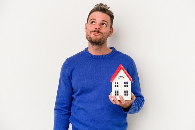 Jonge blanke man met een klein huis geïsoleerd op een witte achtergrond dromen van het bereiken van doelen en doeleinden