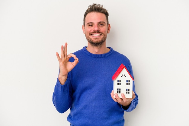 Foto jonge blanke man met een klein huis geïsoleerd op een witte achtergrond vrolijk en zelfverzekerd weergegeven: ok gebaar.