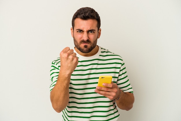 Jonge blanke man met een mobiele telefoon geïsoleerd op een witte achtergrond met vuist naar camera, agressieve gezichtsuitdrukking.