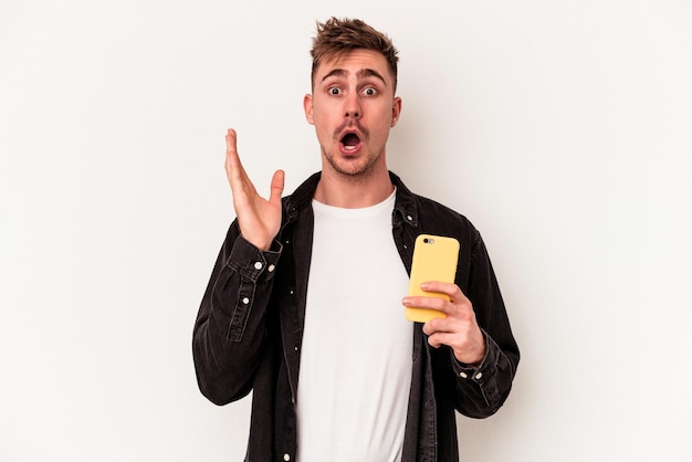 Jonge blanke man met een mobiele telefoon geïsoleerd op een witte achtergrond verrast en geschokt.