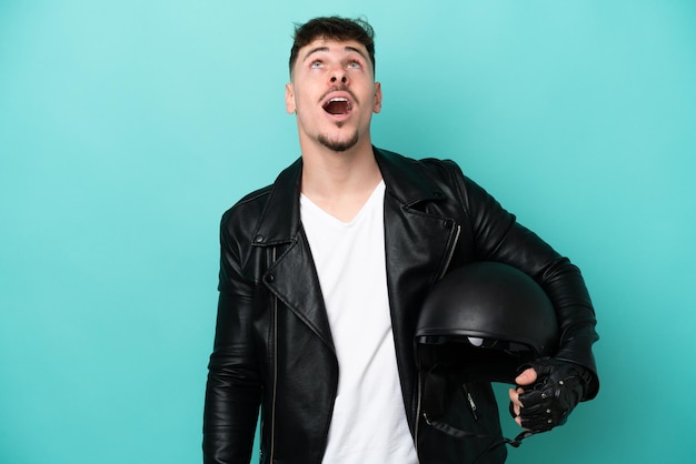 Jonge blanke man met een motorhelm geïsoleerd op blauwe achtergrond opzoeken en met verbaasde uitdrukking