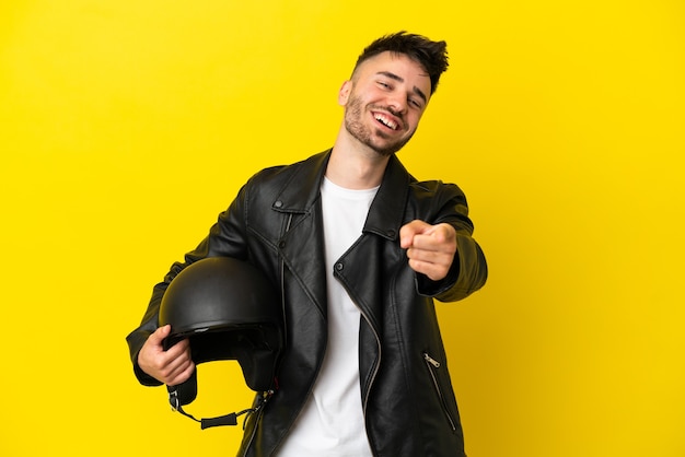 Jonge blanke man met een motorhelm geïsoleerd op een gele achtergrond die naar voren wijst met een gelukkige uitdrukking