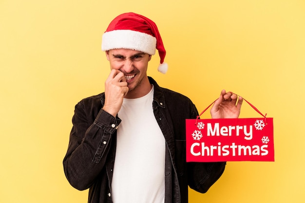 Jonge blanke man met een plakkaat Merry Christmas geïsoleerd op gele achtergrond vingernagels bijten, nerveus en erg angstig.