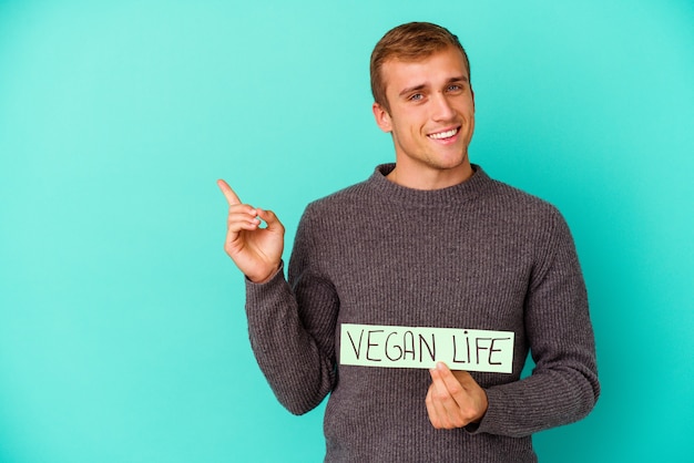 Jonge blanke man met een plakkaat voor veganistisch leven geïsoleerd op een blauwe achtergrond glimlachend vrolijk wijzend met de wijsvinger weg.