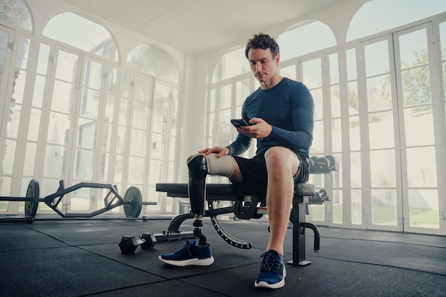 Jonge blanke man met een prosthetisch been zit met een mobiele telefoon op een bankdruk in de sportschool