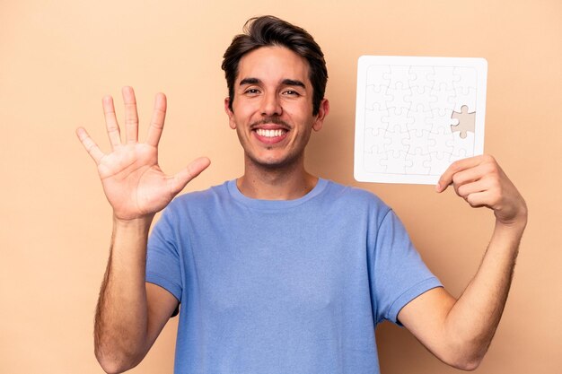 Jonge blanke man met een puzzel geïsoleerd op een beige achtergrond glimlachend vrolijk nummer vijf met vingers.