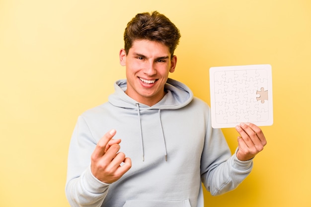 Jonge blanke man met een puzzel geïsoleerd op een gele achtergrond die met de vinger naar je wijst alsof hij je uitnodigt om dichterbij te komen