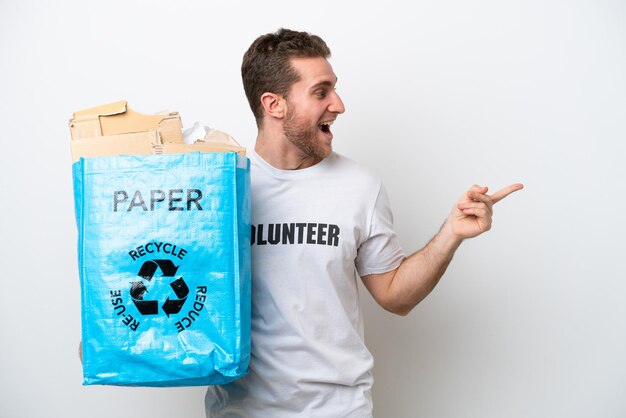 Jonge blanke man met een recyclingzak vol papier om te recyclen geïsoleerd op een witte achtergrond wijzende vinger naar de zijkant en presenteert een product