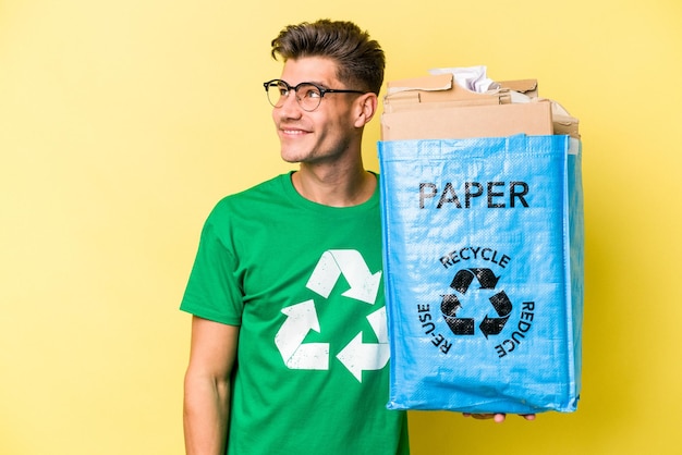 Jonge blanke man met een recyclingzak vol papier om te recyclen geïsoleerd op gele achtergrond, dromend van het bereiken van doelen en doeleinden