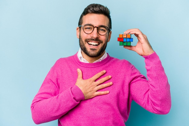 Jonge blanke man met een Rubiks kubus geïsoleerd op blauwe achtergrond lacht hardop terwijl hij zijn hand op zijn borst houdt