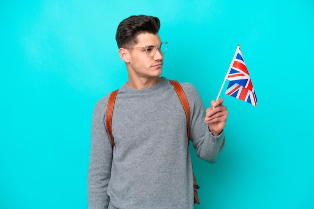 Jonge blanke man met een vlag van het Verenigd Koninkrijk geïsoleerd op een blauwe achtergrond, kijkend naar de zijkant