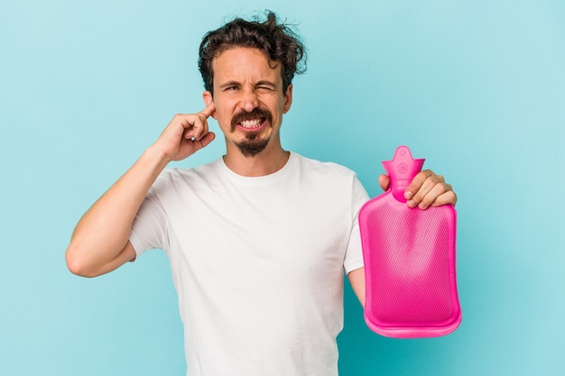 Jonge blanke man met een waterzak geïsoleerd op een blauwe achtergrond die oren bedekt met handen.