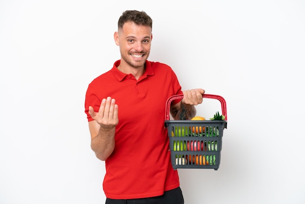 Jonge blanke man met een winkelmandje vol voedsel geïsoleerd op een witte achtergrond die uitnodigt om met de hand te komen Blij dat je gekomen bent