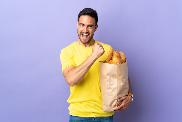 Jonge blanke man met een zak vol brood geïsoleerd op paarse achtergrond een overwinning vieren