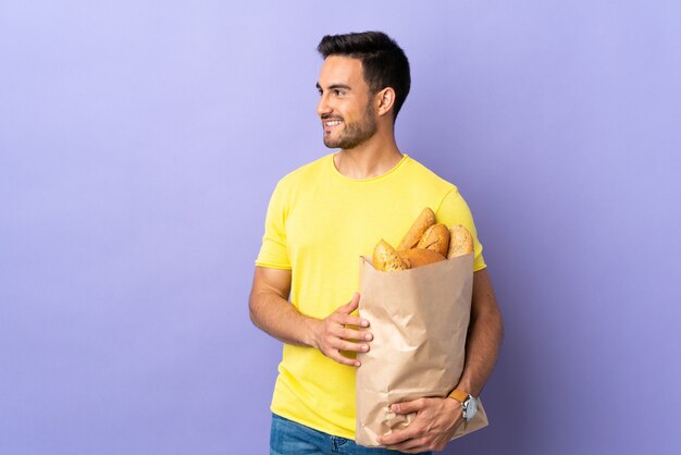 Jonge blanke man met een zak vol brood geïsoleerd op paarse achtergrond op zoek naar de zijkant