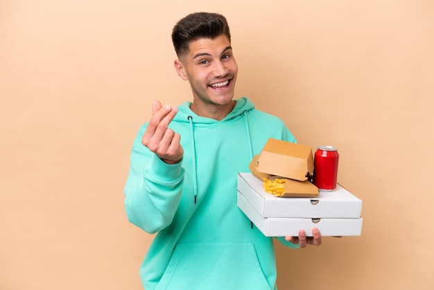 Jonge blanke man met fastfood geïsoleerd op beige achtergrond geld gebaar maken