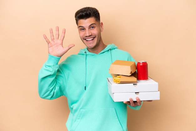 Jonge blanke man met fastfood geïsoleerd op beige achtergrond groetend met hand met gelukkige uitdrukking