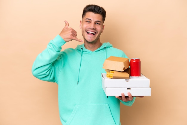 Jonge blanke man met fastfood geïsoleerd op beige achtergrond telefoongebaar maken Bel me terug teken