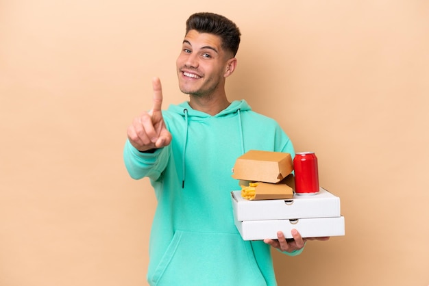 Jonge blanke man met fastfood geïsoleerd op een beige achtergrond die een vinger toont en opheft