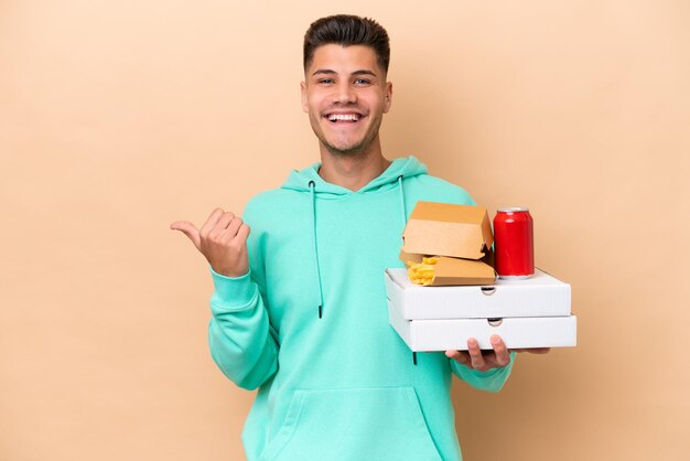 Jonge blanke man met fastfood geïsoleerd op een beige achtergrond, wijzend naar de zijkant om een product te presenteren