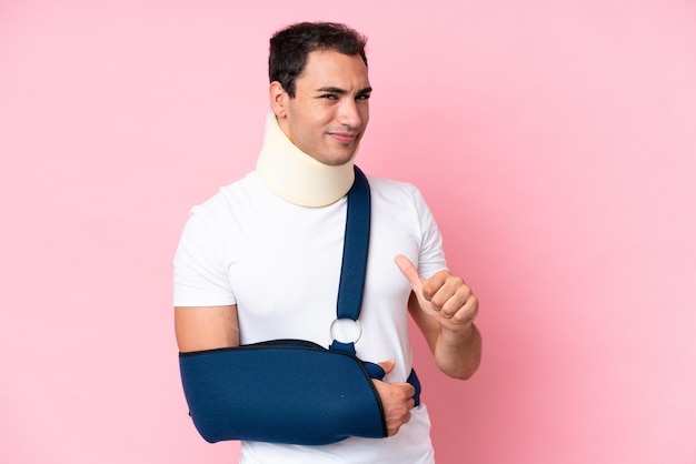 Jonge blanke man met gebroken arm en het dragen van een mitella geïsoleerd op roze achtergrond trots en zelfvoldaan
