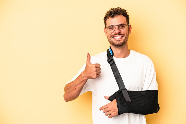 Jonge blanke man met gebroken hand geïsoleerd op gele achtergrond glimlachend en duim omhoog