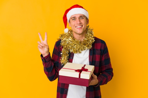 Jonge blanke man met kerstmuts met een cadeau geïsoleerd op gele muur vrolijk en zorgeloos met een vredessymbool met vingers.