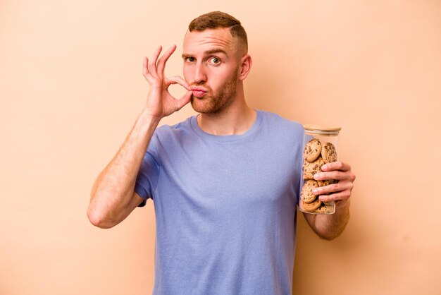 Jonge blanke man met koekjespot geïsoleerd op beige achtergrond met vingers op lippen die een geheim bewaren