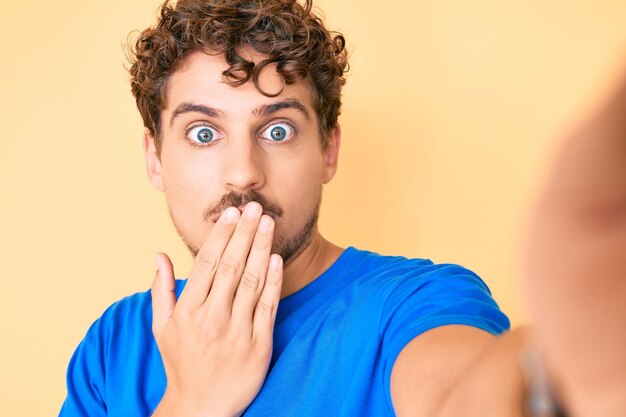 Jonge blanke man met krullend haar, gekleed in vrijetijdskleding die een selfie neemt die de mond bedekt met de hand geschokt en bang voor een fout verraste uitdrukking