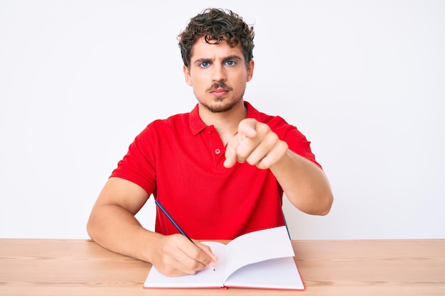 Jonge blanke man met krullend haar schrijvend notitieboekje zittend op de tafel wijzend met vinger naar de camera en naar jou, zelfverzekerd gebaar serieus kijkend