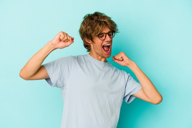 Jonge blanke man met make-up geïsoleerd op blauwe achtergrond die een speciale dag viert, springt en armen met energie opheft.