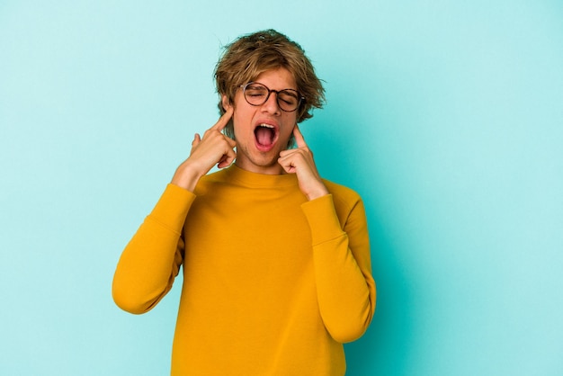 Jonge blanke man met make-up geïsoleerd op blauwe achtergrond die oren bedekt met vingers, gestrest en wanhopig door een luid ambient.