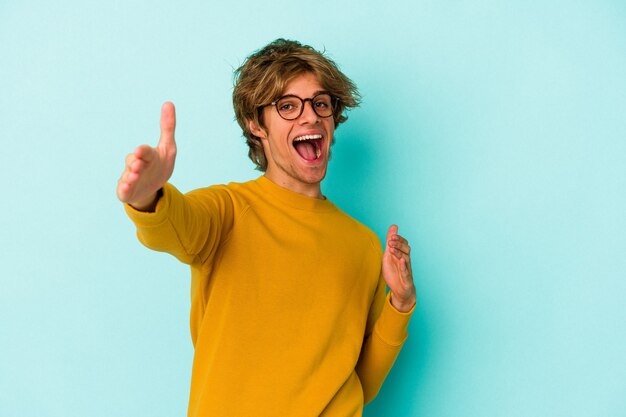 Jonge blanke man met make-up geïsoleerd op blauwe achtergrond voelt zich zelfverzekerd en geeft een knuffel aan de camera.