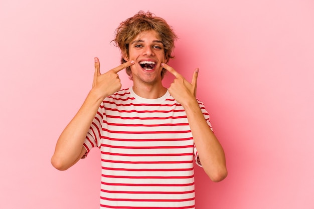 Jonge blanke man met make-up geïsoleerd op roze achtergrond glimlacht, wijzende vingers naar de mond.