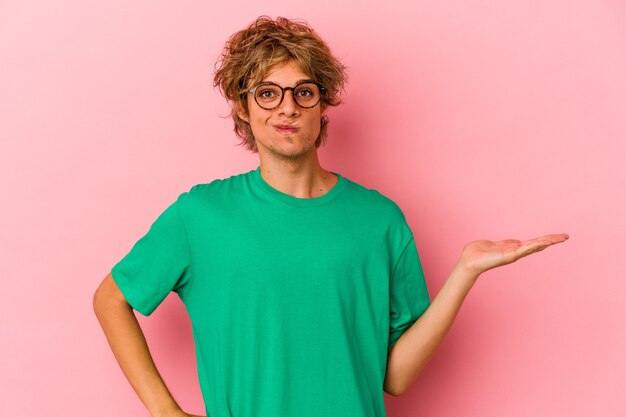 Jonge blanke man met make-up geïsoleerd op roze achtergrond met een kopie ruimte op een handpalm en met een andere hand op de taille.