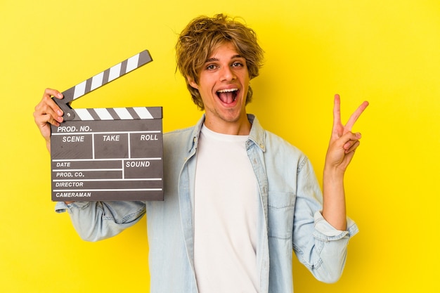 Jonge blanke man met make-up met Filmklapper geïsoleerd op gele achtergrond vrolijk en zorgeloos met een vredessymbool met vingers.