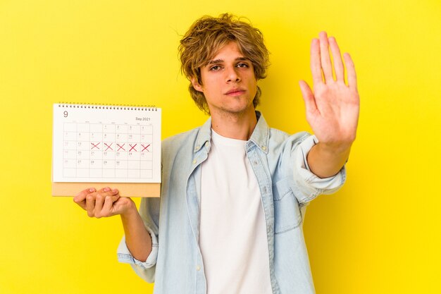 Jonge blanke man met make-up met kalender geïsoleerd op een gele achtergrond, staande met uitgestrekte hand met stopbord, waardoor je wordt voorkomen.