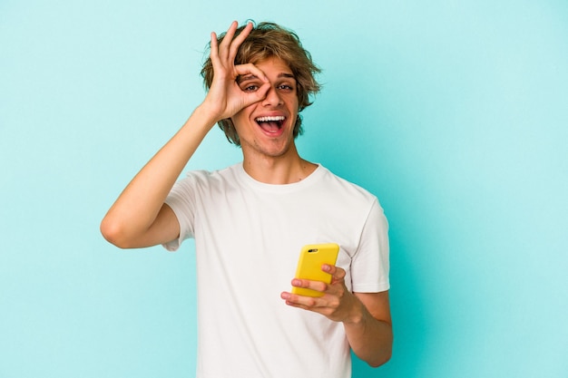 Jonge blanke man met make-up met mobiele telefoon geïsoleerd op blauwe achtergrond opgewonden houden ok gebaar in de gaten.